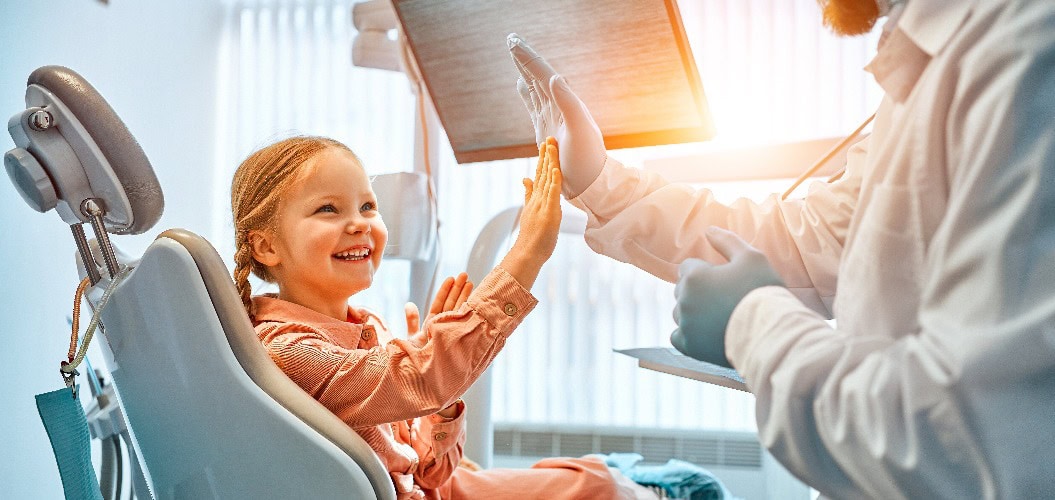 Ein kleines Mädchen sitzt auf einem Zahnarztstuhl, gibt dem Arzt ein High Five und lacht. Zahnpflege, Vertrauen und Patientenbetreuung. Kinderzahnheilkunde.Sonnenlicht.