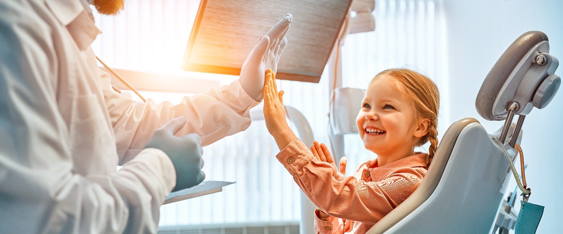 Ein kleines Mädchen sitzt auf einem Zahnarztstuhl, gibt dem Arzt ein High Five und lacht. Zahnpflege, Vertrauen und Patientenbetreuung. Kinderzahnheilkunde.Sonnenlicht.