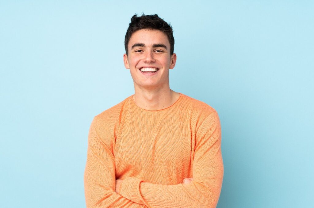 Junger Mann mit dunklen Haaren und orangefarbenem Pullover vor blauem Hintergrund